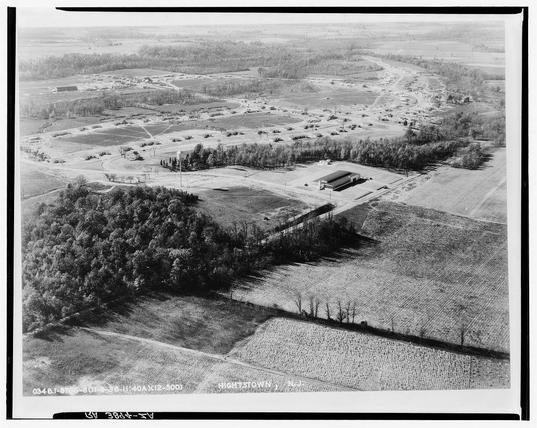 The image is a black and white aerial photograph depicting an expansive landscape with distinct sections of cultivated fields, forests, and built-up areas. In the foreground on the left side lies dense tree-covered land that gradually transitions into open agricultural fields towards the center. The middle ground shows various patches of crops or farmlands arranged in neat rows under clear demarcations. To the right is a larger area with extensive cleared fields extending to the horizon, separated from other sections by more densely wooded areas.

Notable structures are visible within these cultivated regions; one large structure stands prominently on its own field towards the top center, while another substantial building can be seen at the far left edge of the photo. Roads or pathways crisscross through some parts of this landscape, suggesting connectivity between different parcels and buildings. The overall scene suggests an organized agricultural setup with a mix of rural settings.

The photograph is labeled "HIGHTOWN, N.J." indicating that it was taken in Hightstown, New Jersey. There are handwritten inscriptions on the bottom left corner: "034618-5709," and two more lines which seem to be dates or codes, possibly related to cataloging or documentation of this aerial view.

The absence of people in the image focuses attention solely on landscape patterns, suggesting  [...]