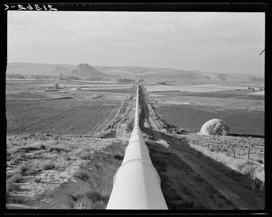 The image depicts a black and white photograph of an expansive rural landscape with a large pipeline stretching from the foreground into the distance. The land appears dry, consisting mostly of open fields with sparse vegetation, indicative of arid or semi-arid climate conditions. In the background lies rolling terrain leading to distant hills. On one side of the field near the center is what looks like a round hay bale covering around 1% of the photo's total area and placed closer than most other objects in view. The pipeline runs vertically through the middle, occupying approximately half of the image from bottom to top.

The photograph exudes an atmosphere of desolation or isolation due to its barren landscape devoid of any visible human activity or structures. It is a monochromatic scene with varying shades of gray and black dominating because it's in grayscale format which gives off vintage vibes reminiscent of early 20th-century photography techniques.