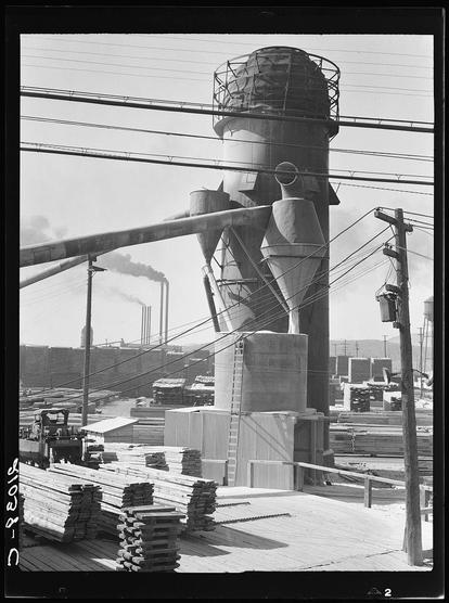 The image captures an industrial scene with a focus on large-scale wood processing equipment. In the foreground, we see stacks of wooden pallets and planks arranged in organized piles. These materials are likely being prepared for use or storage within the facility.

Dominating the background is a towering structure resembling a silo or granary used to store bulk commodities like grain or lumber. This central feature has multiple levels with access points such as ladders, suggesting it's actively utilized and possibly filled with raw material awaiting processing. The silhouette of what appears to be a steam-powered boiler can be seen through openings in the upper tiers.

The environment suggests an active industrial site. Visible smokestacks release dark emissions into the atmosphere, indicative of ongoing operations that generate substantial output or waste products. Power lines crisscross above, reinforcing the sense of a functioning factory zone.

Overall, this black and white photograph likely captures a moment within the early to mid-20th century given its monochromatic quality and style. It encapsulates an era when large-scale lumber processing was prevalent in industrial areas like Klamath Falls, Oregon. The absence of human figures draws attention solely to the machinery's operation and scale.

The image is noted for preserving a historical narrative about industr [...]
