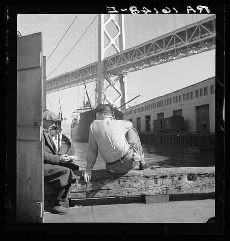 The image depicts two men in a black and white photograph, likely from the early to mid-20th century. One man is sitting on what appears to be a wooden plank or dock with his legs stretched out before him; he's wearing light-colored pants and is holding something small between his fingers that could possibly be food. The second man stands next to him in work attire, including trousers and shoes indicative of manual labor employment, such as those who might have worked at the docks during this era.

In the background, a large industrial bridge with several towers spans across the image, suggesting an urban port setting. A cargo ship is docked near them on calm water under clear skies. The men seem to be taking their lunch break while observing or working alongside maritime activities in San Francisco's waterfront area as indicated by historical context.

The photograph captures a moment of respite and reflection amidst industrial labor with the iconic bridge adding a sense of scale, suggesting an important transportation link during that period.