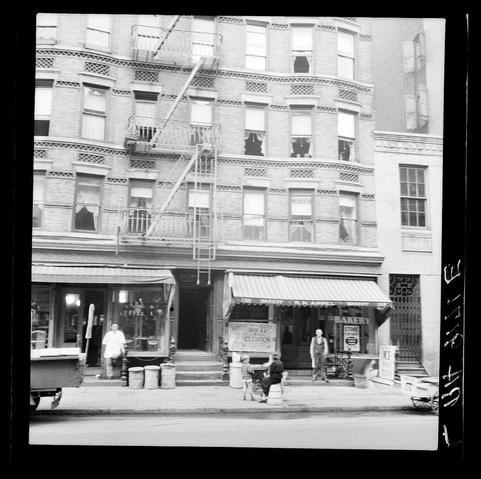 This black and white photograph depicts a street scene from New York City, likely during the early to mid-20th century. The focus is on a large brick building with multiple floors, featuring fire escapes leading up one side of its facade. Several windows are visible; some appear dark while others have curtains drawn or closed blinds.

On the ground floor at the corner appears a bakery and store advertised as "Bakery Store for Rent," indicating commercial activity in the area. A person stands outside on the sidewalk, possibly engaged with work equipment such as bins near them. Another individual is seated closer to the center of the street; it's unclear what they are doing.

A woman sits atop barrels at a lower level than where she likely belongs, suggesting potential neglect or poor working conditions within this tenement-style building. The overall environment appears worn and lived-in with elements like newspapers on stands for public use indicating accessibility and community engagement in daily activities despite apparent hardships implied by the condition of buildings and street setup.

The image conveys a stark portrayal of urban life during its time, emphasizing social issues such as housing conditions, employment struggles, and economic disparity. It captures an intimate moment within this environment that might tell stories about people's lives behind what we see [...]