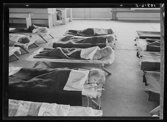The image depicts a group of young children lying on reclining chairs in what appears to be an indoor setting. The room is furnished with rows of empty chairs and benches, suggesting that it could be part of a larger communal space such as a cafeteria or community center. Some beds are covered with blankets while others have only sheets, indicating variations in how the sleeping arrangements were made.

The children appear calm and restful, likely taking advantage of a naptime during their day at school. The overall atmosphere is peaceful, despite being filled with numerous individuals within close proximity to each other.