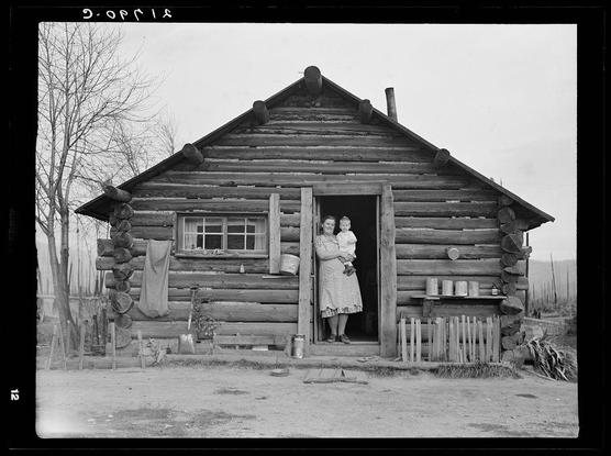 The image depicts a vintage black and white photograph of an old, rustic log cabin. A woman stands in the doorway with her arm around one child while holding another baby on her hip. The wooden structure appears aged, constructed from horizontal logs tied together at right angles where they meet the corners. Various items are hung or propped against the wall outside, including what seem to be a hat and other personal belongings, suggesting domestic life in rural living conditions.

The background is relatively barren with leafless trees and an open sky visible above. The ground appears dry and uneven around the cabin's foundation. There’s also some sort of stack of items on the right side near the door that looks like chopped wood or perhaps dried crops.

There are handwritten markings at the top left corner, possibly indicating a date "9-Oct-13," but it could be misread as such because they're not clear in this format. The bottom-right part contains another marking which reads '2,' likely representing page number two of an album or collection within a larger set.

From additional information provided about the image (not shown here), it seems to mention that this log house is now occupied and has been enlarged by the Halley family with Mrs. Halley standing in the doorway, possibly indicating her role as matriarch during those times. It suggests a historical setting or co [...]