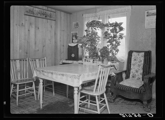 The image depicts a vintage black and white photograph of an interior room, likely from the early to mid-20th century. The setting appears to be a dining area within a home with wooden paneling on the walls. A table covered with a floral-patterned cloth is positioned at the center, surrounded by four chairs: two plain wooden side chairs, one ornate upholstered armchair, and another simple chair.

On top of the table rests an old radio and some potted plants near the window, which allows natural light to enter through sheer curtains. To the right stands a decorative plant in a pot with visible soil layers. The floor is covered by patterned linoleum or carpet tiles. On one wall hangs framed artwork or photographs, while another shows a calendar indicating it's January.

The overall ambiance of the room suggests a modest yet cozy domestic space from an earlier era, reflecting simplicity and functionality typical for that period in residential design.