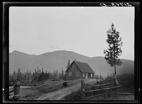 The image depicts a rural landscape with a small wooden house in the foreground. The structure appears aged, suggesting it might be of historical interest or significant to the area's history. It is situated on what looks like farmland surrounded by fencing and sparse vegetation typical of arid climates. Behind the house stands a tall evergreen tree that contrasts against an expansive mountain range under an overcast sky.

A noticeable sign indicates "FOR SALE," hinting at potential changes in land ownership or property development. The black-and-white tone adds to the nostalgic atmosphere, evoking themes related to rural life and possibly historical preservation efforts for farming communities like those mentioned in Boundary County, Idaho.