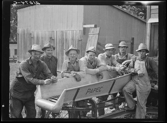 The image depicts a group of seven farmers wearing overalls, hats, and protective eyewear as they pose with their co-operatively owned ensilage cutter. They are standing on what appears to be farm equipment near the silo filled with ensilage for animals' feed during winter season in Oregon. The ensilage cutter is labeled "PAPEC," indicating it's a brand or model of farming machinery used at that time period, possibly in 1940s given by its vintage style and grayscale photo quality.